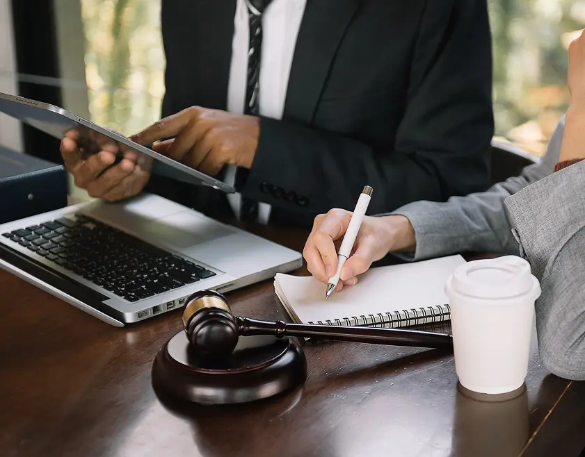Two office workers working together, one holds a tablet and the other writes on a notebook.